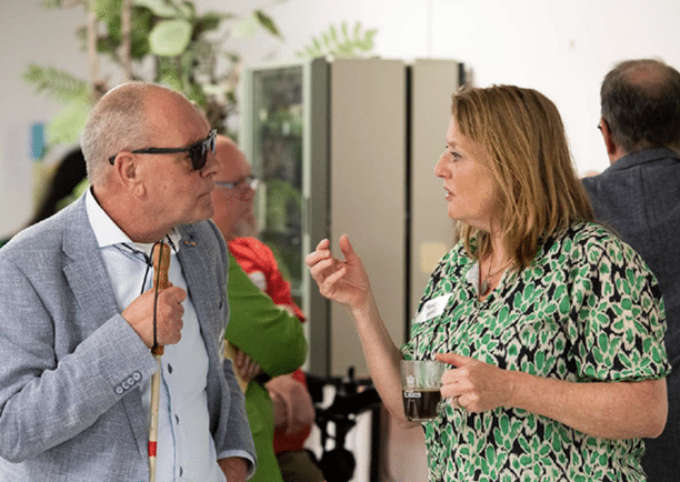 Een man met een zonnebril en blindenstok praat op een bijeenkomst met een vrouw