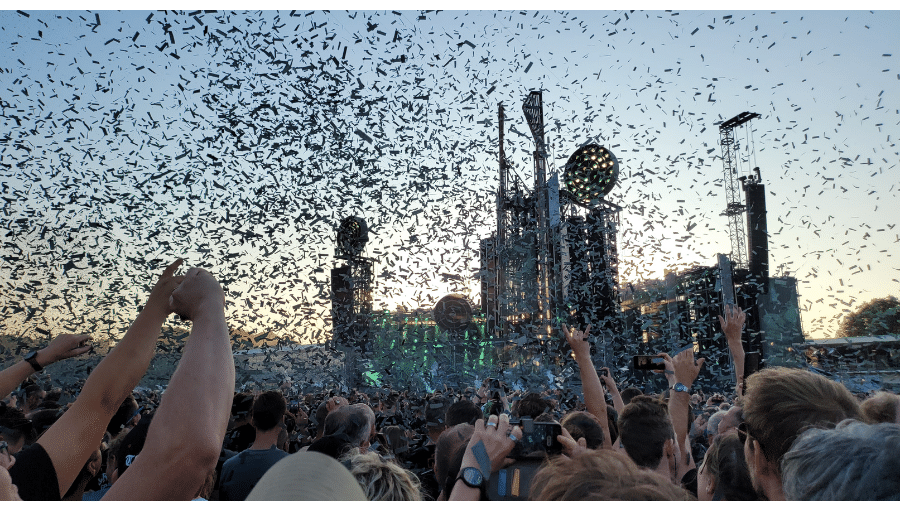een grote mensenmassa kijkt naar een immens podium van rammstein. er vliegt confetti door de lucht. een vrouw maar een hartjesgebaar met haar handen