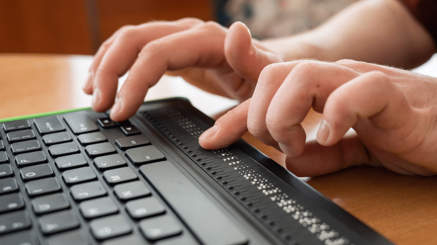 een hand typt op een toestenbord van een laptop terwijl de andere hand een brailleleesregel aan het voelen is