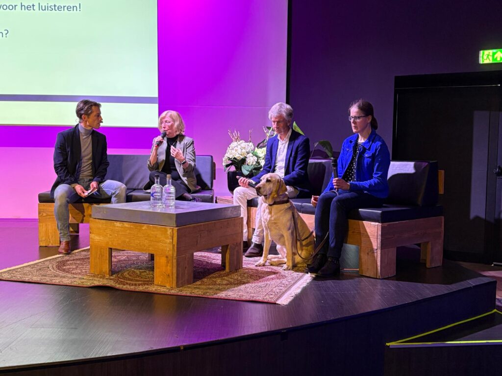 vier sprekers zitten op het podium op bankjes te discussieren, twee oogartsen, dagvoorzitter Niels Crama en Mirjam Boers met haar geleidehond Chimo voeren het gesprek