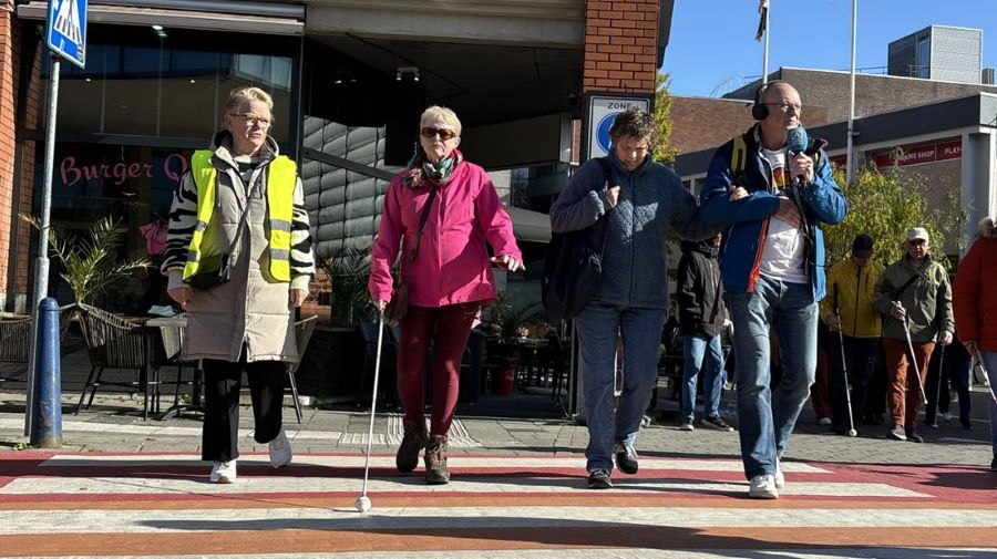 Twee mensen met een visuele beperking, een begeleider en journalist steken een zebrapad over