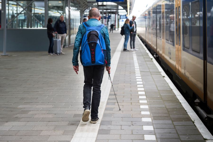 Een man met een witte stok volgt de geleidelijn op een perron