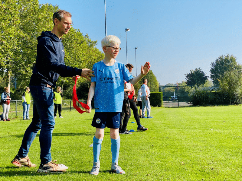 een buurtsportcoach staat achter een jongentje met albinisme aan de rand van een sportveld. De sportcoach lijkt hem een voorzichtig maar bemoedigend duwtje in de rug te geven om het veld in te gaan. De jongen draagt een sportshirt van Manchester City, heeft wit haar en draagt een bril, hij lijkt te zwaaien naar iemand op het veld