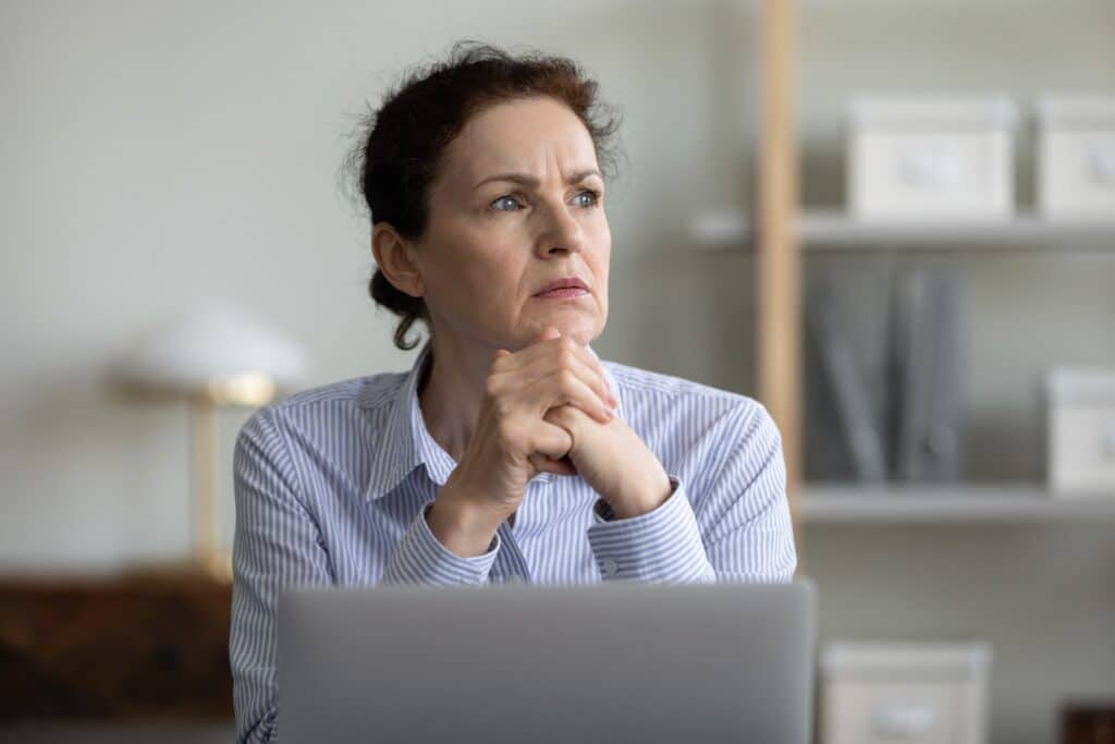 een vrouw met bruin haar en een blouse aan zit te mijmeren achter haar laptop. ze kijkt onzeker en twijfelachtig weg van haar laptop terwijl ze knijpt in haar handen samen