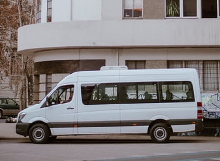 een wit taxibusje wacht langs de stoep, er kunnen ongeveer 8 tot 10 personen in.