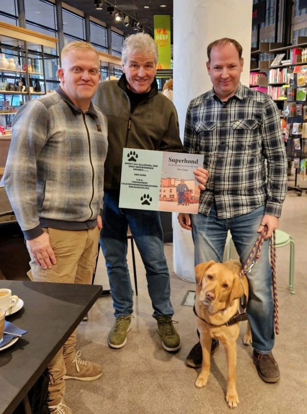 In het midden Kees Tinga met de cheque van €500 in zijn hand, de cheque ziet eruit als de kaft van het boek Superhond. Links van Kees staat Maikel en rechts staat Geert met geleidehond Abby, allen kijken naar de camera.